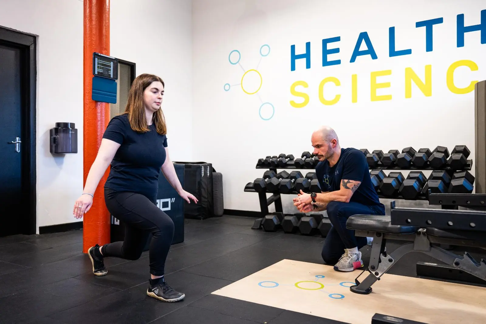 KA3_2759 Female athlete performing a reverse lunge with a personal trainer in a gym setting.