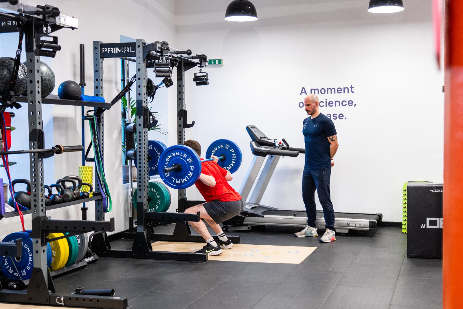 KA3_3107 A man performing a back squat with a barbell while a personal trainer edinburgh observes his form at Health By Science Leith.