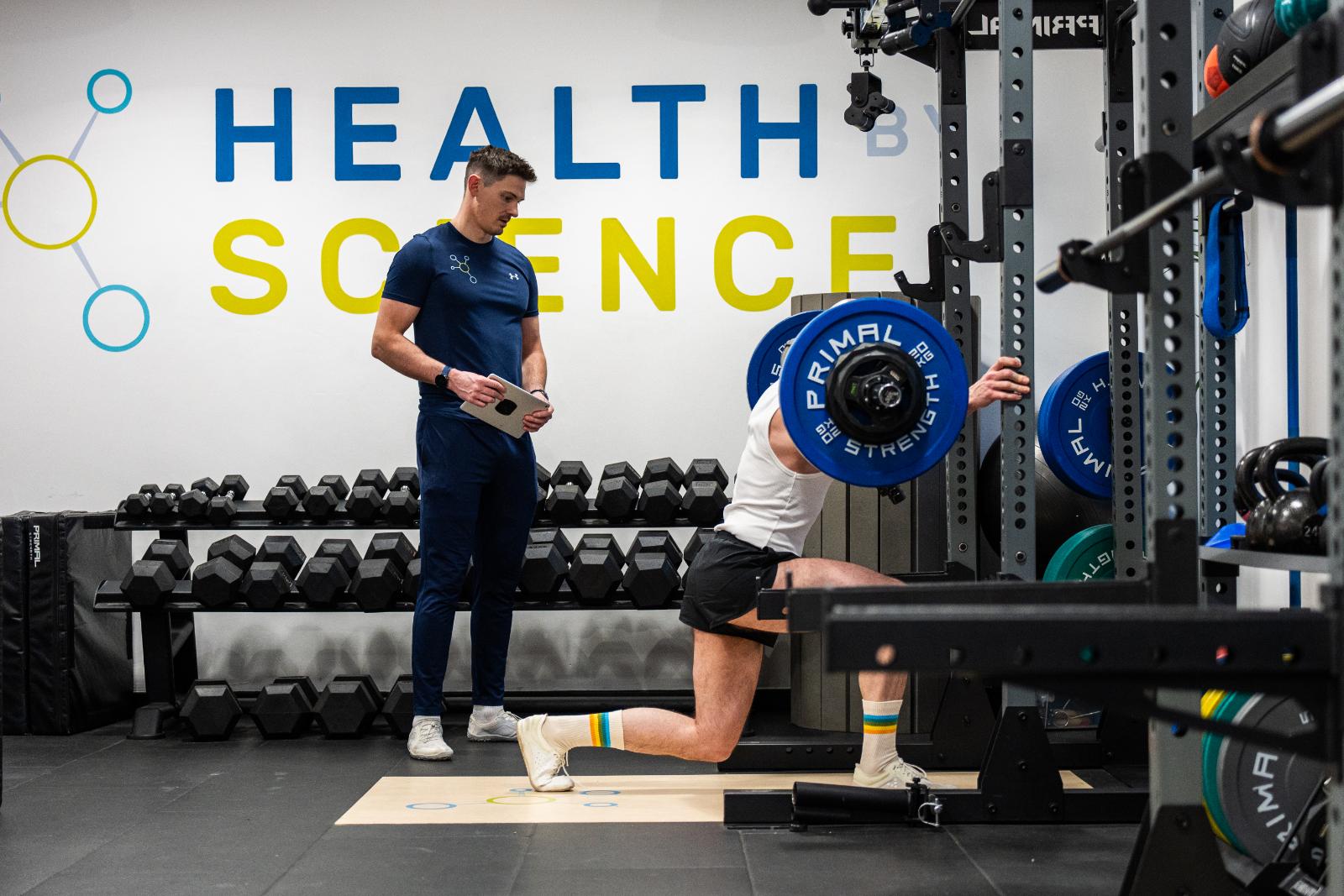 KA3_3703 Health by Science coach monitoring a barbell split squat to assess unilateral leg strength and knee stability.